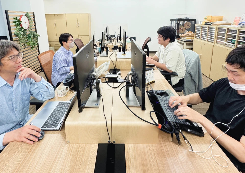 写真:社内の作業風景。向かい合ってデスク作業する4名のスタッフ
